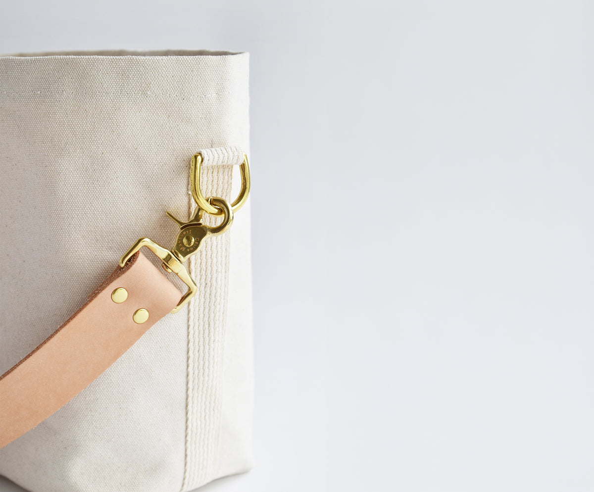 Close-up of a canvas tote bag corner with a light tan leather strap attached via gold-tone hardware against a plain white background.