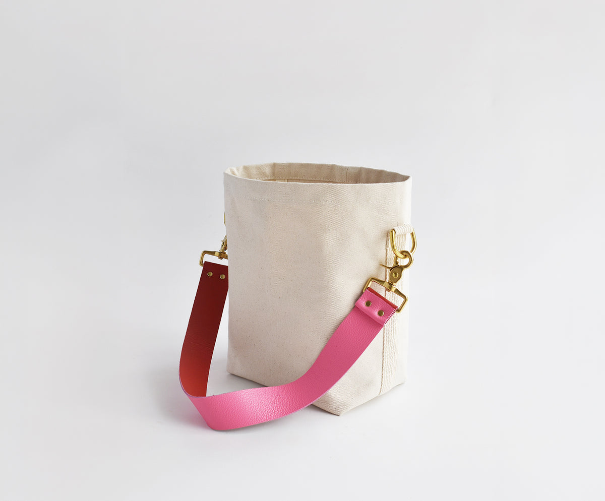 A canvas bucket bag in a light beige color, featuring a bright pink adjustable shoulder strap and a red accent strap, both attached with gold-tone hardware, set against a plain light gray background.