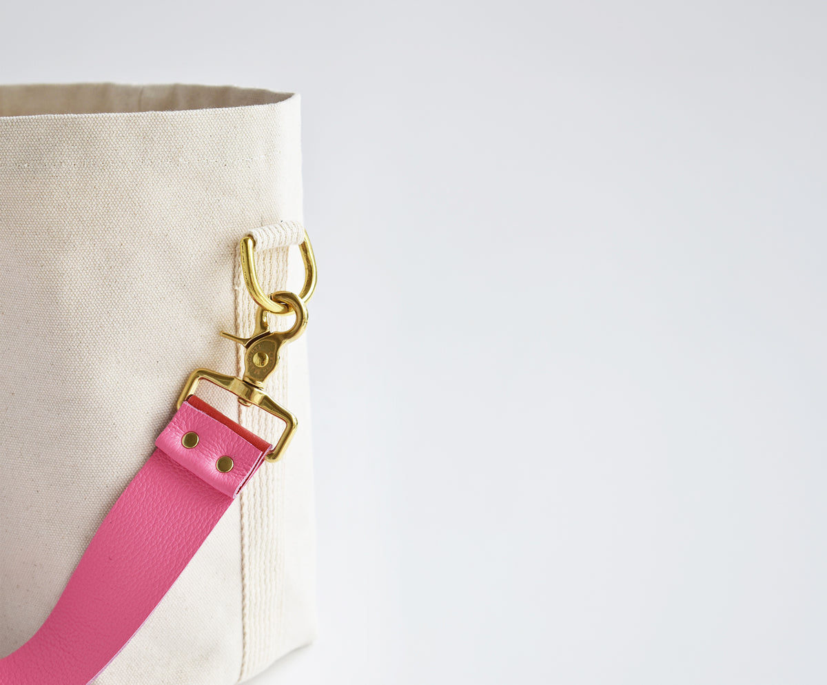 Close-up of a canvas tote bag in off-white with a bright pink leather strap and gold-tone hardware on a plain light background.