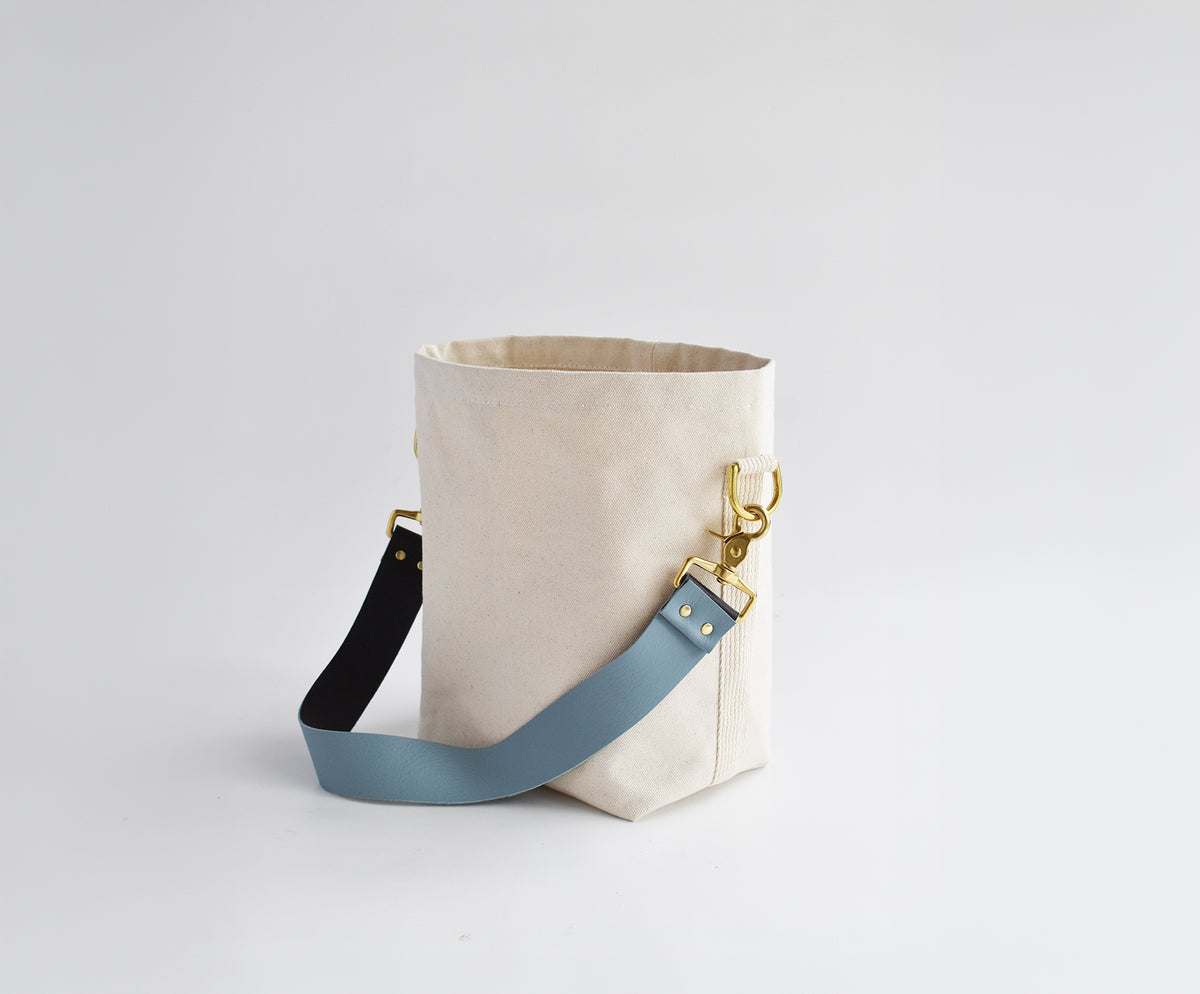 A canvas bucket bag in natural beige with a light blue leather shoulder strap, gold-tone hardware, and a black accent section on the strap, photographed on a seamless light gray background.