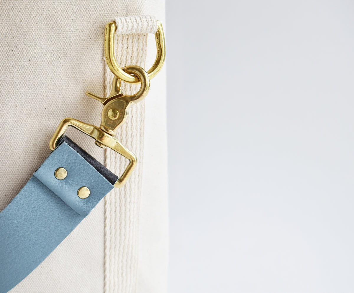 Close-up of a light blue leather strap with gold-tone hardware attached to a cream canvas bag, set against a clean white background.