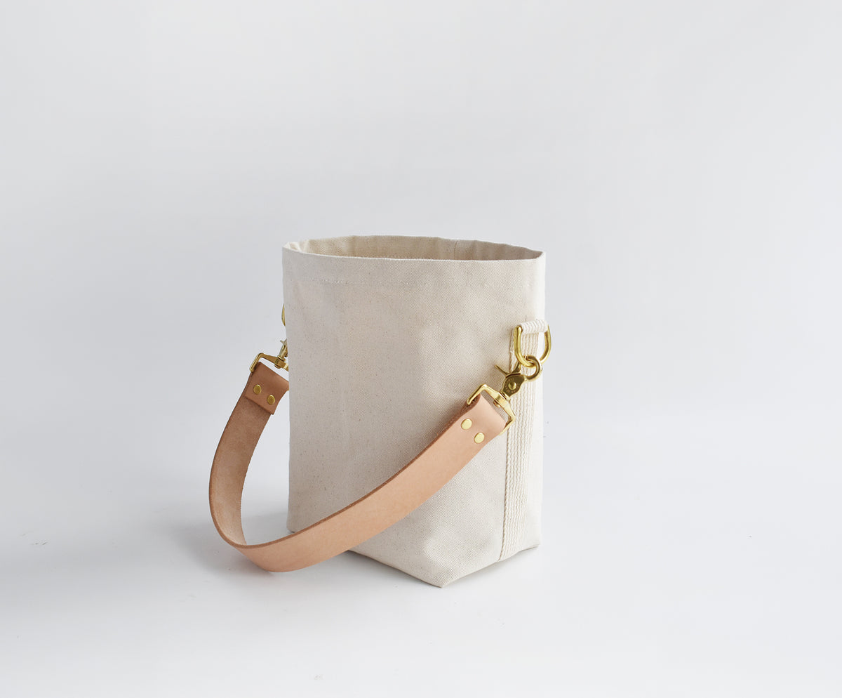 A light beige canvas bucket bag with a tan leather shoulder strap and gold-tone metal hardware, photographed on a seamless light gray background.